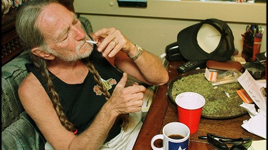 American country singer Willie Nelson takes a drag off a joint while relaxing at his home in Texas, 2000s. A large amount of marijuana is spread out on the table before him (Photo by Liaison/Getty Images)